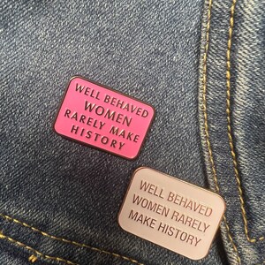 May include: Two enamel pins with the text "Well Behaved Women Rarely Make History". The top pin is pink with gold text. The bottom pin is white with gold text.