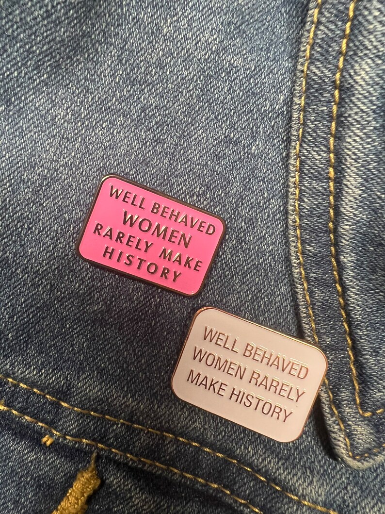 May include: Two enamel pins with the text "Well Behaved Women Rarely Make History". The top pin is pink with gold text. The bottom pin is white with gold text.