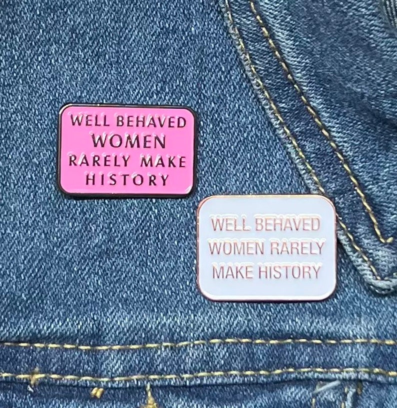 May include: Two enamel pins with the text "Well Behaved Women Rarely Make History". The top pin is pink with black text. The bottom pin is white with rose gold text.