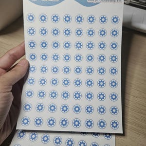 May include: Two sheets of white sticker paper with blue and white star-shaped stickers. The stickers are arranged in a grid pattern. The top sheet has the text "Plush Planning" and a website address.
