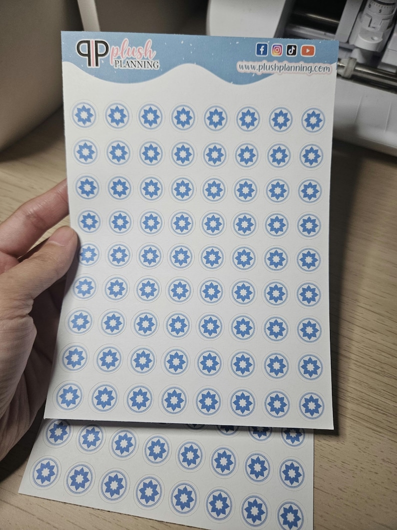 May include: Two sheets of white sticker paper with blue and white star-shaped stickers. The stickers are arranged in a grid pattern. The top sheet has the text "Plush Planning" and a website address.