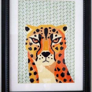 May include: A framed cross-stitch artwork featuring a close-up of a cheetah's face. The cheetah is depicted in orange, black, and white against a green background.