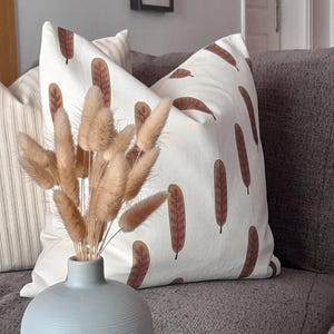 以下が含まれることがあります： 茶色の葉柄の白いクッション、縦縞のベージュのクッション、そして茶色のソファの上に置かれた、ドライパンプキンを飾った花瓶
