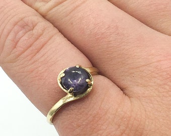 May include: A gold ring with a purple gemstone set in a prong setting. The ring has a curved band.