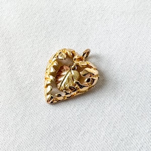 May include: A gold-colored heart-shaped pendant with a textured border and a leaf design in the center. The pendant is small and has a loop at the top for a chain. The background is a white fabric.