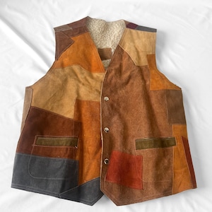 May include: A sleeveless vest constructed from various patches of suede leather in shades of brown, tan, orange, and grey. The garment features a cream-colored sherpa lining, a V-neckline, and three silver-toned metal snap buttons down the front.