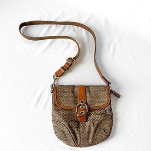 May include: Brown Coach crossbody bag with a brown leather strap and accents. The bag features a repeating pattern of the Coach logo in a lighter brown color. The bag has a flap closure with a buckle and a brown leather strap.