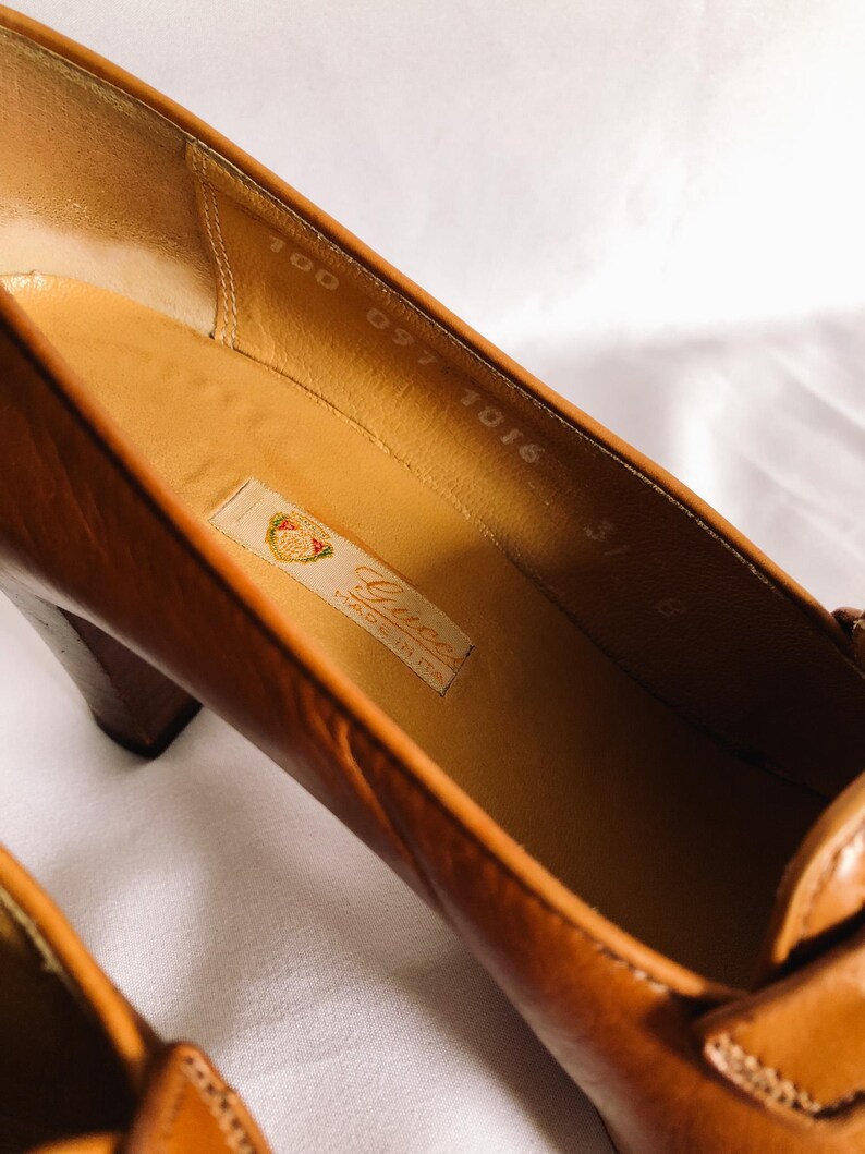 May include: Close-up of a pair of brown leather high heels. The interior of the shoe is a light tan colour, with a label that reads "Gucci Made in Italy." The heel is a dark brown colour.
