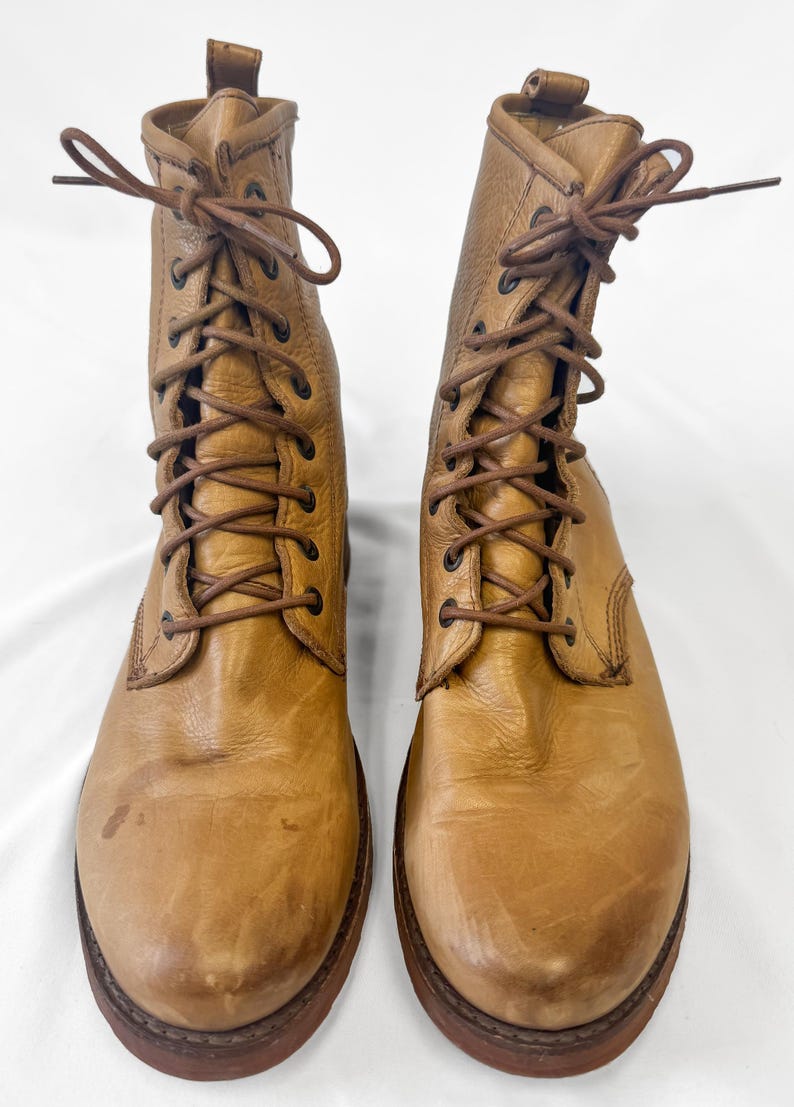 May include: A pair of tan leather lace-up boots. The boots have a worn appearance with visible creases and scuffs. The soles are dark brown, and the laces are a matching brown color. The boots are displayed against a white background.