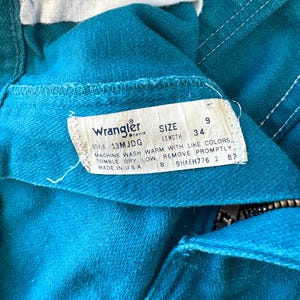 May include: A teal denim garment featuring a white interior tag with the Wrangler brand logo, size 9, and length 34. The tag includes care instructions: machine wash warm with like colors, tumble dry low, remove promptly, and made in USA.