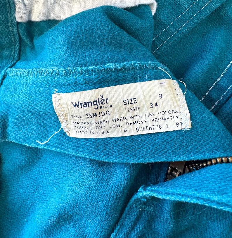 May include: A teal denim garment featuring a white interior tag with the Wrangler brand logo, size 9, and length 34. The tag includes care instructions: machine wash warm with like colors, tumble dry low, remove promptly, and made in USA.