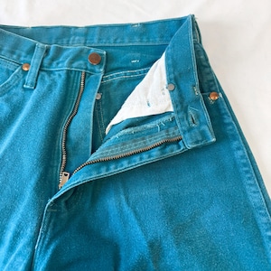 May include: A pair of teal denim jeans featuring a high-waisted design with a metal zipper fly, a button closure at the waistband, and copper-toned rivets at the pocket corners. The interior pocket lining is white fabric, and the seams are stitched.