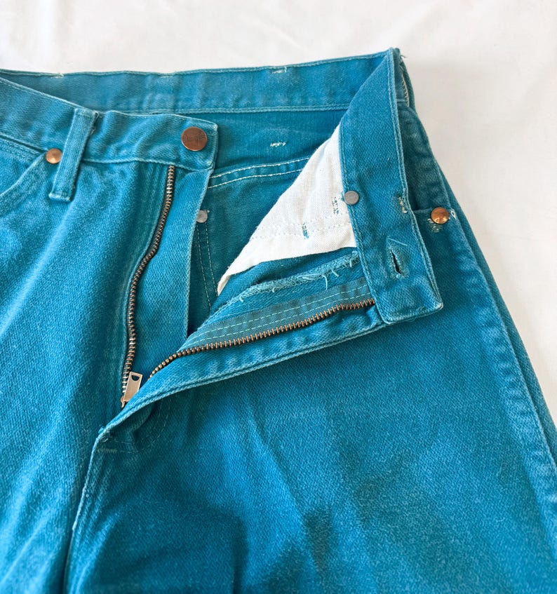 May include: A pair of teal denim jeans featuring a high-waisted design with a metal zipper fly, a button closure at the waistband, and copper-toned rivets at the pocket corners. The interior pocket lining is white fabric, and the seams are stitched.