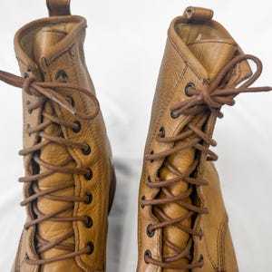 May include: A pair of tan leather lace-up boots. The boots have a textured surface, brown laces, and metal eyelets. The boots are shown against a white background.