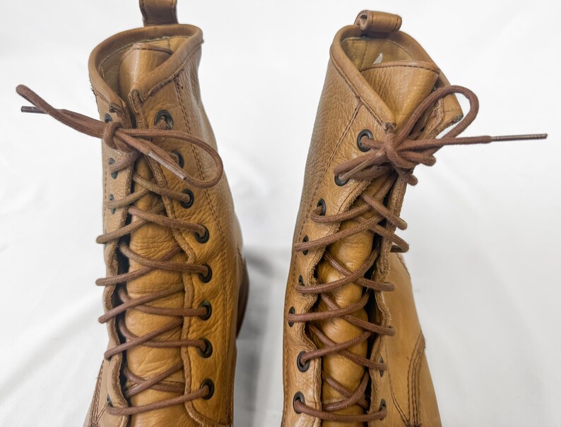 May include: A pair of tan leather lace-up boots. The boots have a textured surface, brown laces, and metal eyelets. The boots are shown against a white background.