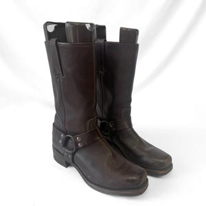 May include: A pair of dark brown leather boots with a square toe and a harness detail. The boots have a mid-calf height and a side zipper. The leather has a textured appearance. The boots are on a white background.