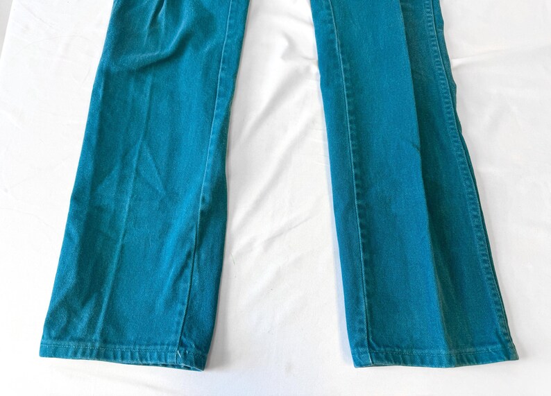 May include: A pair of teal-colored pants featuring a straight-leg cut and a classic denim-style construction with visible vertical seams along the legs. The fabric has a consistent solid teal hue throughout, with a standard hem at the bottom.