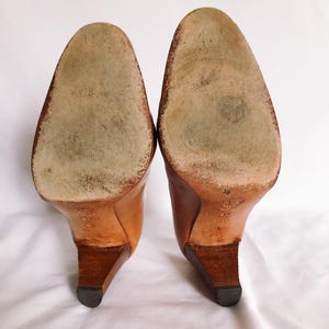 May include: A pair of light brown leather high-heeled shoes, seen from the soles. The heels are a darker brown, and the soles show wear. The shoes are set against a white background.