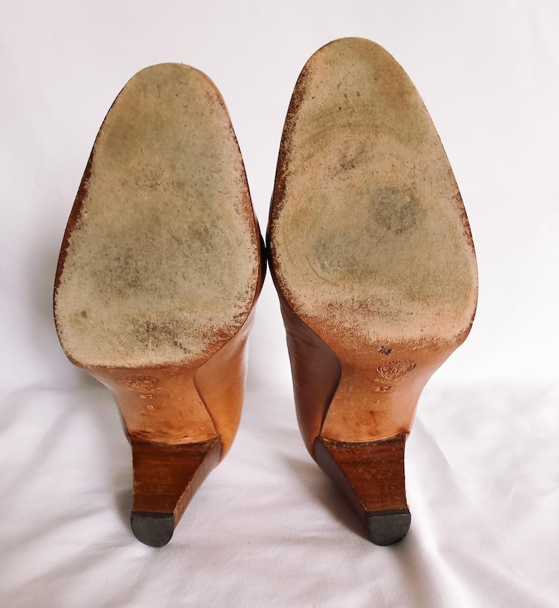 May include: A pair of light brown leather high-heeled shoes, seen from the soles. The heels are a darker brown, and the soles show wear. The shoes are set against a white background.