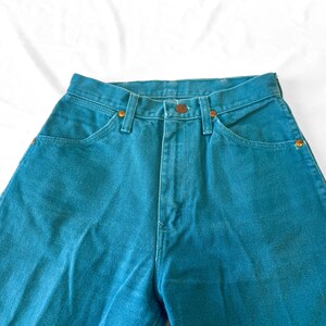 May include: High-waisted teal denim jeans featuring a classic five-pocket design, a front zipper fly with a button closure, and copper-toned metal rivets at the pocket corners. The fabric has a solid, saturated teal color throughout the garment.