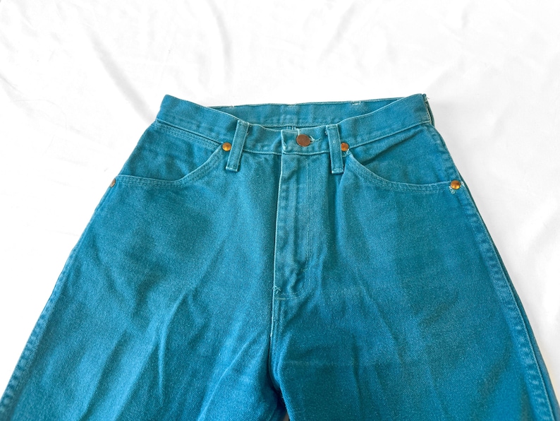 May include: High-waisted teal denim jeans featuring a classic five-pocket design, a front zipper fly with a button closure, and copper-toned metal rivets at the pocket corners. The fabric has a solid, saturated teal color throughout the garment.