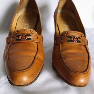 May include: A pair of tan leather heeled loafers. Each shoe has a strap across the vamp with a gold-toned buckle and green and red accents. The shoes have a rounded toe and a low heel.