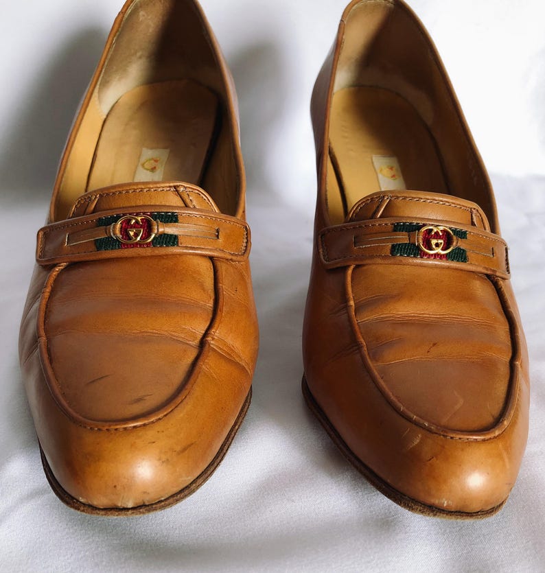 May include: A pair of tan leather heeled loafers. Each shoe has a strap across the vamp with a gold-toned buckle and green and red accents. The shoes have a rounded toe and a low heel.