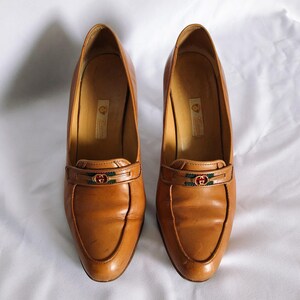 May include: A pair of tan leather heeled loafers. Each shoe has a decorative strap with a green and red emblem. The shoes have a pointed toe and a small heel. The shoes are displayed on a white surface.