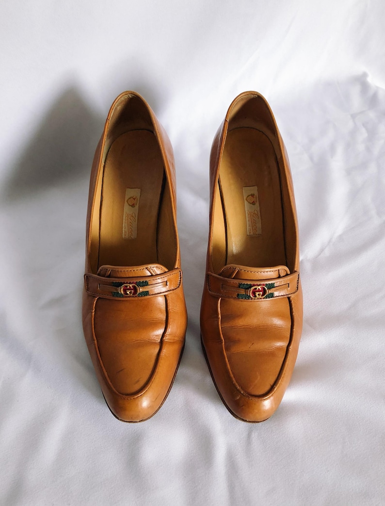 May include: A pair of tan leather heeled loafers. Each shoe has a decorative strap with a green and red emblem. The shoes have a pointed toe and a small heel. The shoes are displayed on a white surface.