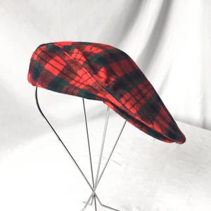 May include: A red and green plaid newsboy cap. The cap is displayed on a metal stand against a white background. The plaid pattern features a bold red color with dark green accents, creating a classic and stylish look. The cap's design is a traditional flat cap style.