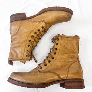 May include: Two tan leather lace-up boots with brown soles and heels. The boots have a worn, vintage appearance, with visible creases and scuffs. The laces are brown, and the boots are set against a white background.
