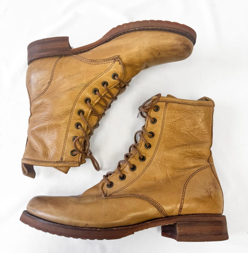 May include: Two tan leather lace-up boots with brown soles and heels. The boots have a worn, vintage appearance, with visible creases and scuffs. The laces are brown, and the boots are set against a white background.