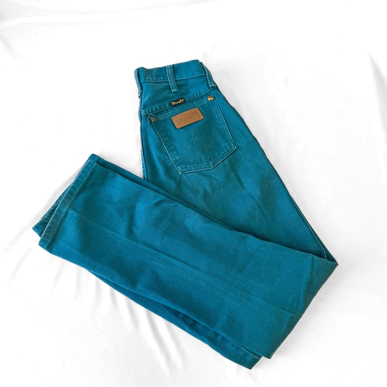 May include: Teal-colored denim jeans featuring a high-waisted design, a single back pocket with a tan leather patch, a metal button closure, and belt loops. The Wrangler brand logo is visible on the leather patch and a small black tag.