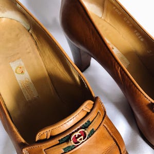 May include: Close-up of two brown leather Gucci pumps. The shoes have a block heel and a decorative strap with the Gucci logo in gold, red, and green. The shoes are made in Italy.