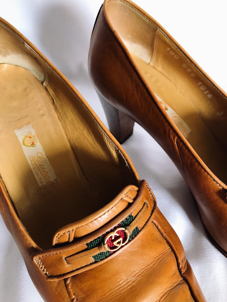 May include: Close-up of two brown leather Gucci pumps. The shoes have a block heel and a decorative strap with the Gucci logo in gold, red, and green. The shoes are made in Italy.