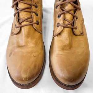 May include: A pair of tan leather boots with brown laces. The boots have a worn appearance with visible creases and scuffs. The soles are dark brown, and the boots are designed with a lace-up closure. The boots are a classic style.