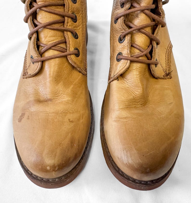 May include: A pair of tan leather boots with brown laces. The boots have a worn appearance with visible creases and scuffs. The soles are dark brown, and the boots are designed with a lace-up closure. The boots are a classic style.