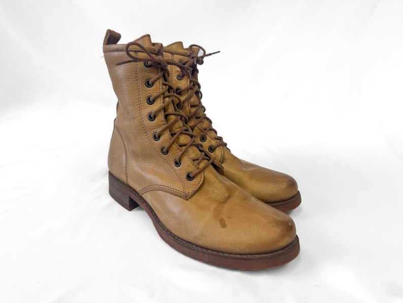 May include: A pair of tan leather combat boots with brown laces. The boots have a rounded toe and a dark brown sole. The leather shows signs of wear, giving them a vintage look. The boots are approximately 8 inches tall.