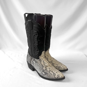 May include: A pair of black and cream-colored cowboy boots featuring a textured snakeskin-patterned leather foot and a smooth black leather shaft with decorative stitching. The boots have a pointed toe, a stacked heel, and pull tabs at the top.