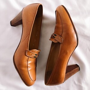 May include: A pair of tan leather heeled loafers. The shoes have a rounded toe, a strap across the top with a decorative buckle, and a wooden block heel. The shoes are set against a white background.