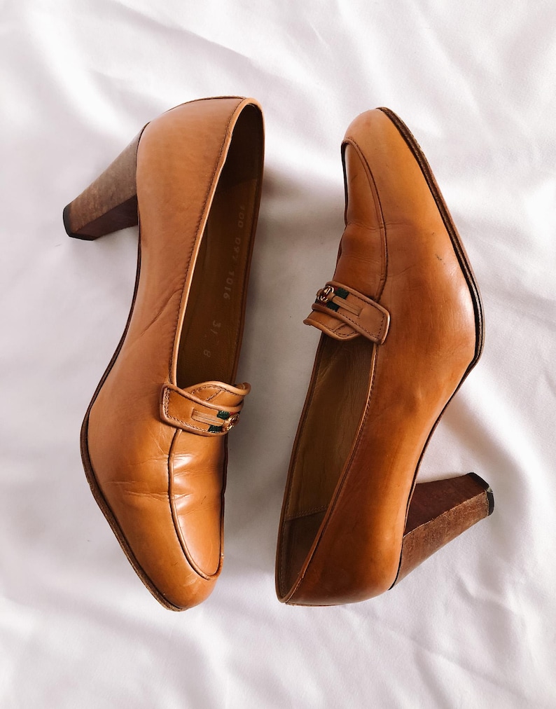 May include: A pair of tan leather heeled loafers. The shoes have a rounded toe, a strap across the top with a decorative buckle, and a wooden block heel. The shoes are set against a white background.