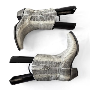 May include: A pair of mid-calf boots featuring a grey and white snakeskin pattern. The boots have a pointed toe, a stacked heel, and black plastic boot trees inserted inside to maintain their shape. The design includes vertical paneling.