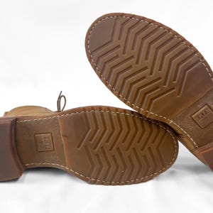 May include: Close-up of the soles of two brown leather boots. The soles have a textured pattern and are stitched to the boot. The heels are a darker brown. The brand name is visible on the sole.