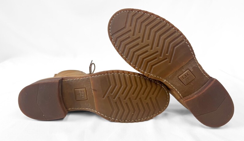 May include: Close-up of the soles of two brown leather boots. The soles have a textured pattern and are stitched to the boot. The heels are a darker brown. The brand name is visible on the sole.