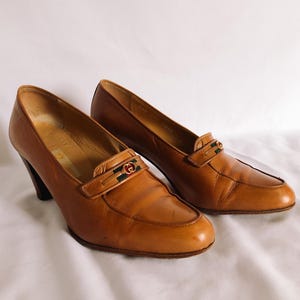 May include: A pair of brown leather heeled loafers. Each shoe has a decorative strap with a gold-tone buckle and green and red accents. The heels are dark brown and the shoes are set against a white background.