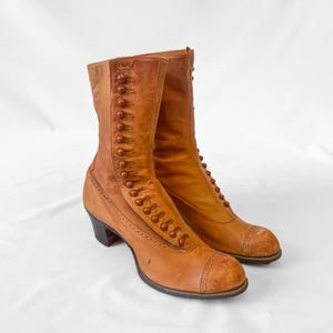 May include: A pair of tan leather mid-calf boots featuring a row of decorative buttons along the side, a rounded toe with perforated detailing, and a dark brown block heel. The boots have a structured shaft and a smooth finish throughout.