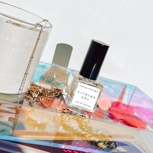 May include: A clear glass bottle of perfume with a black cap. The bottle is labeled "Gold Palms Citrine Sol Eau de Parfum". The bottle is sitting on a iridescent tray with gold jewelry and a pink and red plastic object.
