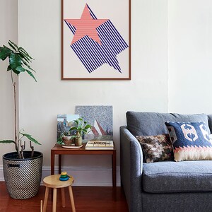 K&ouml;nnte beinhalten: Ein gerahmter Druck eines roten und blauen Sternmusters mit der Zahl 1776 in der oberen linken Ecke. Der Druck h&auml;ngt an einer wei&szlig;en Wand &uuml;ber einem grauen Sofa mit Kissen.