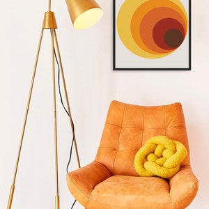 May include: A gold tripod floor lamp with a white lampshade sits next to a brown leather armchair with a yellow knitted throw pillow. A framed print with the text "BAUHAUS" and a geometric design of concentric circles in shades of orange, yellow, and brown hangs on the wall.