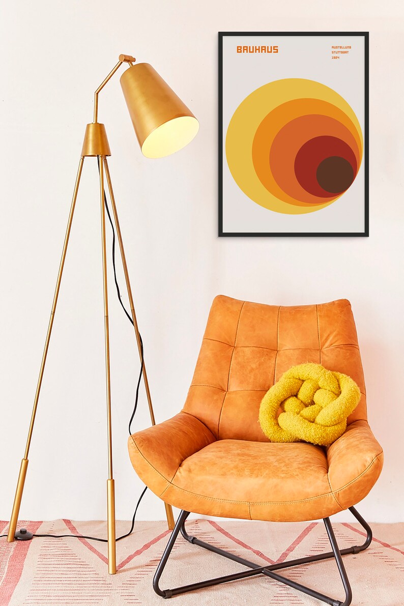 May include: A gold tripod floor lamp with a white lampshade sits next to a brown leather armchair with a yellow knitted throw pillow. A framed print with the text "BAUHAUS" and a geometric design of concentric circles in shades of orange, yellow, and brown hangs on the wall.